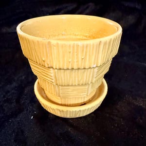 May include: A pale yellow ceramic plant pot with a matching saucer, featuring a tiered, basket-weave texture on the exterior. The pot has a wide, open top and a tapered base, with a glossy finish that shows subtle crackle patterns across the surface.