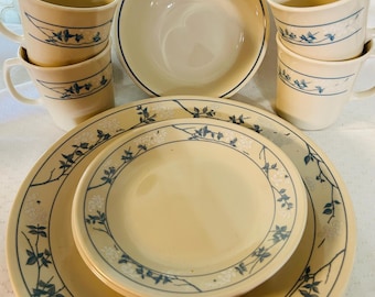 May include: A set of Corelle dinnerware in the "Winter Frost" pattern. The set includes a large dinner plate, a salad plate, a bowl, and four mugs. The plates and bowl feature a white background with a blue and white floral design. The mugs have a cream background with a blue and white floral design.