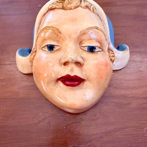 May include: A ceramic wall plaque shaped like a human face wearing a light-colored head covering with blue accents. The face features painted blue eyes, rosy cheeks, and bright red lips, with a glossy finish and a smooth, rounded facial structure.