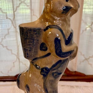 May include: A tan ceramic rabbit figurine with blue painted details, standing upright on a circular base. The rabbit features a basket-like structure on its back and stylized markings on its body, with a glossy finish and upright ears.