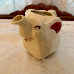 May include: A cream-colored ceramic creamer shaped like an elephant. The pitcher features a trunk that serves as a spout, a handle on the back, and painted red accents on the ears. The elephant has small brown painted eyes and a curved mouth.