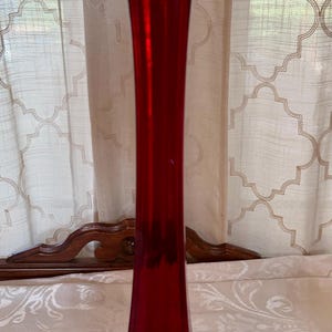 May include: A tall, ruby-red glass vase with a flared base and a ruffled rim. The vase is approximately 12 inches tall and has a slender, elongated shape. The background includes a patterned curtain and a decorative wooden piece.
