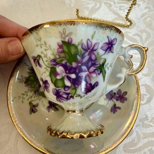 May include: A vintage teacup and saucer set. The teacup and saucer are white with a gold rim and a floral design featuring purple violets and green leaves. The teacup has a pedestal base and a gold handle. The saucer has a matching floral design.