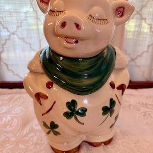 May include: A ceramic cookie jar shaped like a pig with a cream-colored body, a dark green scarf around its neck, and green shamrock patterns on its belly. The pig has closed eyes with painted eyelashes, a pink snout, and brown-tipped hooves.