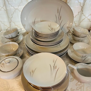 May include: A complete white porcelain dinnerware set with a wheat stalk design and silver trim. The set includes plates, bowls, cups, saucers, a serving platter, a gravy boat, and a covered serving dish. The dishes are arranged on a white tablecloth.