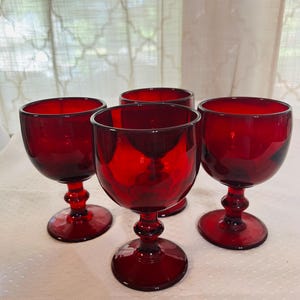 May include: Four ruby red wine glasses. Each glass has a rounded bowl, a decorative stem, and a circular base. The glasses are made of a transparent material, allowing light to pass through, and are arranged on a white surface.
