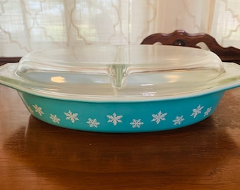 May include: A vintage turquoise Pyrex divided casserole dish with a clear glass lid. The dish features a snowflake pattern around the rim. The dish is on a wooden table.