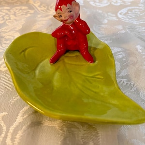 May include: A whimsical ceramic figurine of a red elf perched on a bright green leaf-shaped dish. The elf has a smiling face and is wearing a pointed hat. The dish has a glossy finish, adding to the charm of this decorative piece.