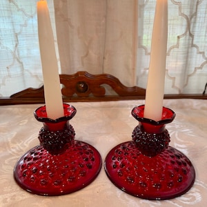May include: Two red glass candle holders with white candles. The candle holders have a hobnail design and are lit with a flame. The candle holders are on a white surface.