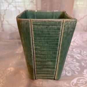 May include: A square, light green ceramic vase with a textured surface. The vase has a trapezoidal shape with vertical, horizontal, and angled lines. The vase is a decorative item.