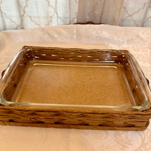 May include: A rectangular glass baking dish with a woven brown wicker basket. The glass dish is empty and sits inside the basket. The basket has a rectangular shape with a woven pattern. The background is a light-colored tablecloth.