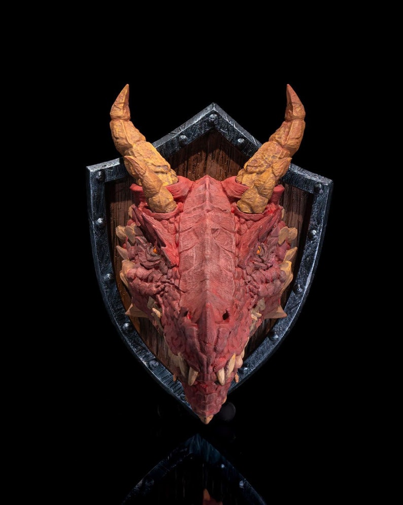 Puede incluir: Una cabeza de drag&oacute;n roja montada en un escudo de madera con un borde de metal negro. El drag&oacute;n tiene cuernos amarillos y dientes afilados.