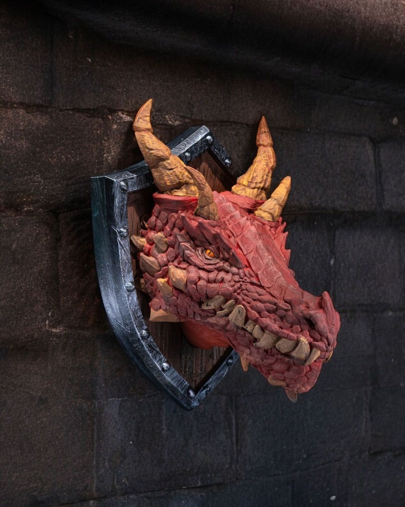 Puede incluir: Una cabeza de drag&oacute;n roja montada en un escudo de madera con un borde de metal plateado. El drag&oacute;n tiene una piel texturizada, escamosa y dientes afilados.