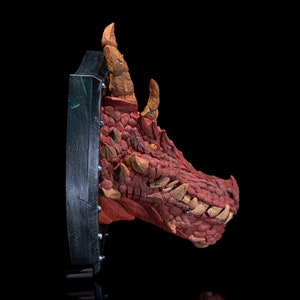 Puede incluir: Una escultura de cabeza de drag&oacute;n roja montada sobre una base circular negra y plateada. El drag&oacute;n tiene una expresi&oacute;n feroz con dientes afilados y una piel texturizada y escamosa.