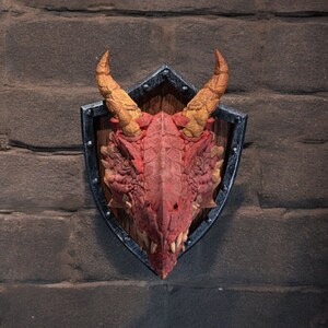 Puede incluir: Una cabeza de drag&oacute;n roja montada en un escudo negro y plateado con un fondo de madera. El drag&oacute;n tiene cuernos amarillos y una expresi&oacute;n feroz.