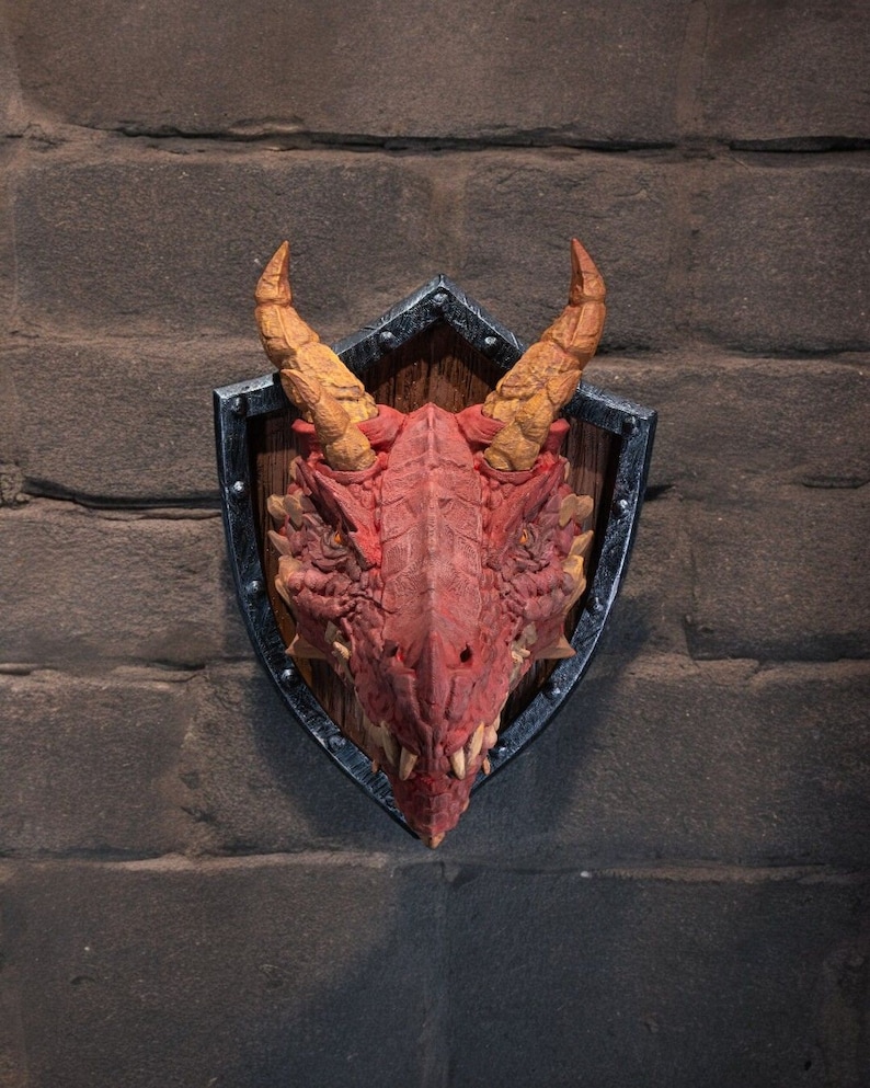 Puede incluir: Una cabeza de drag&oacute;n roja montada en un escudo negro y plateado con un fondo de madera. El drag&oacute;n tiene cuernos amarillos y una expresi&oacute;n feroz.