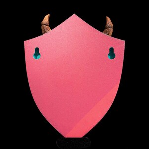 Puede incluir: Un soporte de pared de color rosa en forma de escudo con dos cuernos marrones en la parte superior y dos agujeros de colgar de color turquesa. El soporte est&aacute; hecho de madera y tiene un acabado liso y brillante.