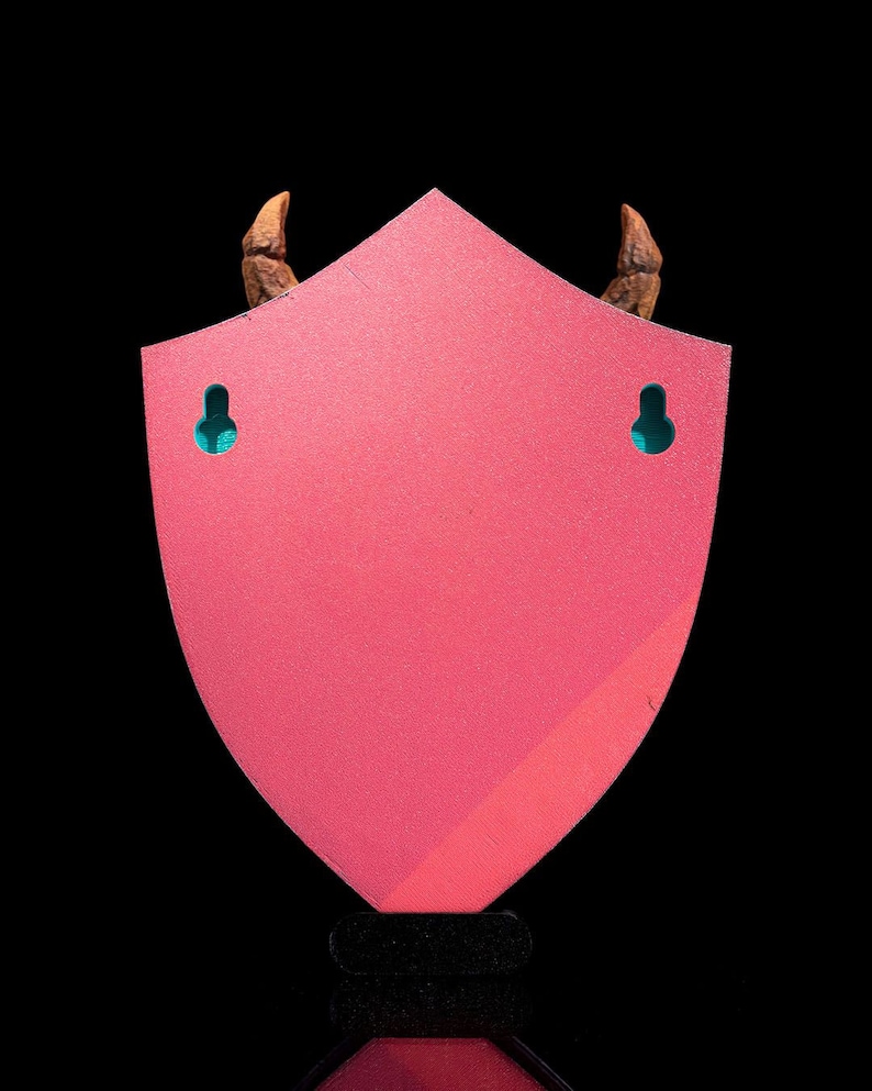 Puede incluir: Un soporte de pared de color rosa en forma de escudo con dos cuernos marrones en la parte superior y dos agujeros de colgar de color turquesa. El soporte est&aacute; hecho de madera y tiene un acabado liso y brillante.