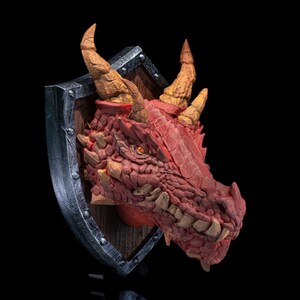 Puede incluir: Una escultura de cabeza de drag&oacute;n roja montada en un escudo. El drag&oacute;n tiene una piel texturizada y escamosa y dientes afilados. El escudo es de metal y tiene un respaldo de madera.