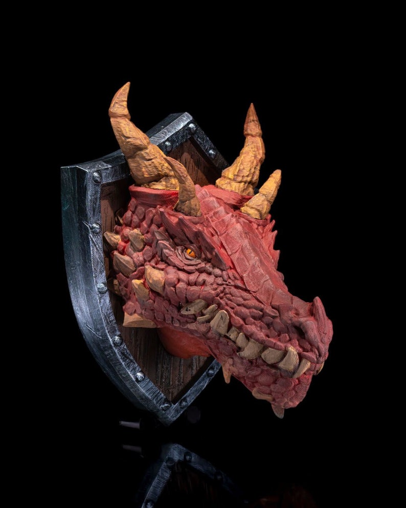 Puede incluir: Una escultura de cabeza de drag&oacute;n roja montada en un escudo. El drag&oacute;n tiene una piel texturizada y escamosa y dientes afilados. El escudo es de metal y tiene un respaldo de madera.