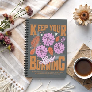 May include: A gray spiral bound notebook with the text "Keep Your Light Burning" in orange and white lettering. The notebook has a floral design with pink flowers and orange leaves. The text "For where your treasure is, there your heart will be also. And where your heart is, there your treasure will be also. Luke 12:34-35" is printed in white at the bottom of the notebook.