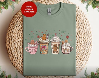 Puede incluir: Camiseta verde salvia con un diseño navideño con cuatro hombres de jengibre en tazas de chocolate caliente con crema batida y bastones de caramelo. El texto "MÁS COLORES DISPONIBLES" está en un círculo rojo en la esquina superior izquierda.