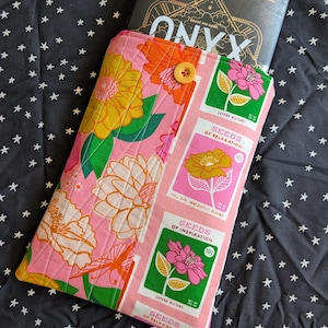May include: A pink, orange, and yellow floral patterned book sleeve with a button closure. The sleeve contains a book titled "ONYX". The design incorporates seed packet graphics with the text "SEEDS OF RELAXATION" and "SEEDS OF INSPIRATION".