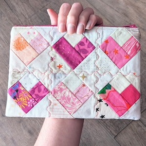 May include: A quilted pouch with a patchwork design. The pouch features a variety of pink and white fabrics, arranged in a geometric pattern. The design includes heart shapes and small star and floral accents. A pink zipper secures the top.