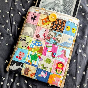 以下が含まれることがあります： 様々な動物やキャラクターのデザインが施された、カラフルなパッチワーク生地の電子書籍リーダーケース。 ケースはボタン留め式で、電子書籍リーダーを収納できます。 背景は白い星模様のダークファブリックです。