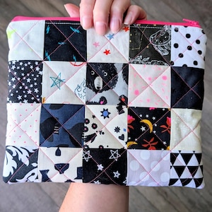 May include: A rectangular quilted zipper pouch featuring a patchwork design of various black, white, and gray fabric squares. The patterns include stars, polka dots, geometric shapes, and animal motifs, all stitched together with a bright pink thread.