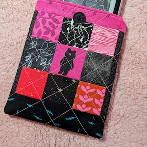 May include: A quilted e-reader case with a patchwork design. The case features a variety of fabrics in pink, black, and red, with a black bear silhouette on a pink square. A black button secures the flap. The case is designed to hold an e-reader.