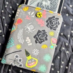 以下が含まれることがあります： 花、クモ、ポーションボトルのカラフルな模様が施されたグレーの電子書籍リーダーケース。ケースには、本の表紙「A Fate」が表示された電子書籍リーダーが入っています。黄色のボタンでケースを閉じます。