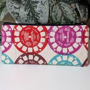 May include: A rectangular pencil case with a black zipper. The case is patterned with colorful, circular designs in red, purple, teal, and brown, resembling vintage film reels. The background is a natural, off-white fabric.