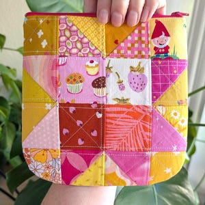 以下が含まれることがあります： 様々な色の生地の正方形を使ったパッチワークのファスナー付きポーチ。正方形には、カップケーキ、イチゴ、ノームの模様があります。ポーチはピンク色のファスナー付きで、手で持たれています。