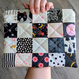 May include: A rectangular quilted pouch featuring a patchwork design of various fabric squares. The patterns include a black bear, stars, moons, flowers, polka dots, stripes, and abstract shapes in a color palette of black, white, and pink.