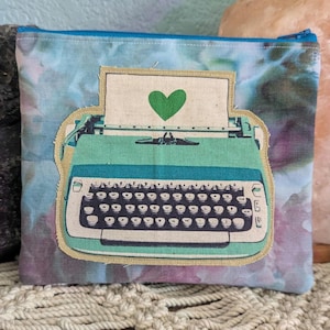 Może przedstawiać: Saszetka zapinana na zamek z tłem w stylu akwareli w odcieniach niebieskiego, fioletowego i różowego. Saszetka posiada naszywkę z zabytkową miętową maszyną do pisania z papierem z zielonym sercem. Zamek błyskawiczny jest niebieski.