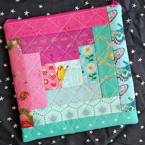 May include: A quilted fabric pouch with a pink zipper. The patchwork design features pink, teal, and white fabrics with butterfly, floral, and crown patterns. The pouch is quilted with a diamond pattern.
