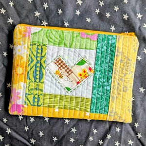 May include: A colorful, quilted zipper pouch with a patchwork design. The pouch features a heart-shaped appliqué in the center, surrounded by various patterned fabrics in shades of yellow, green, and pink. The zipper is yellow.
