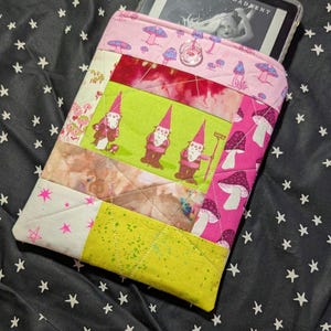 Op de afbeelding: Een gewatteerde e-reader hoes met een patchwork ontwerp. De hoes heeft roze, groene en gele panelen met paddenstoel- en kabouterillustraties. De e-reader toont de tekst "CARISSA BROADENT".