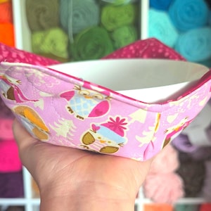 Puede incluir: Un protector de cuenco rosa con un diseño de búhos y árboles. Sostiene un cuenco blanco. El fondo muestra estantes llenos de madejas de hilo de colores. El protector está diseñado para mantener los cuencos calientes.