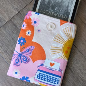 May include: A colorful e-reader case with a floral and butterfly pattern. The case features a pink, orange, and white design with a sun, flowers, and a typewriter graphic. The e-reader is visible inside the case, displaying the title "ALCHEMISED".