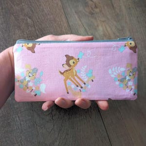 Puede incluir: Una bolsa rectangular rosa con cremallera con un patrón repetido de un ciervo de dibujos animados, flores y otros elementos del bosque. La bolsa tiene una cremallera gris y se sostiene en una mano.