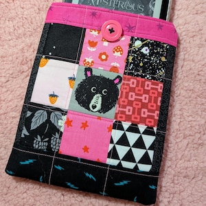 May include: A patchwork e-reader sleeve with a variety of fabric patterns, including strawberries, mushrooms, stars, and a bear. The sleeve is secured with a pink button. An e-reader with the title "Mysterious" is visible inside the sleeve.
