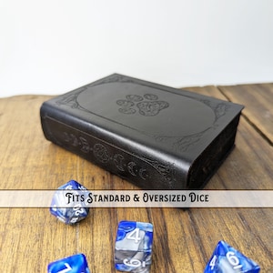Peut inclure: Un coffret à dés noir en cuir avec un motif de patte de chien en relief sur le couvercle. Le coffret rectangulaire est posé sur une surface en bois avec des dés bleus et blancs. Le texte sur l'image indique : FITS STANDARD & OVERSIZED DICE.