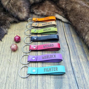 May include: Seven leather keychains in orange, tan, lime green, black, red, purple, and turquoise, each featuring a silver metal ring and a snap button. The keychains are embossed with the words BARD, WARLOCK, ROGUE, BARBARIAN, WIZARD, WARLOCK, and FIGHTER.