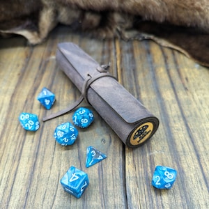 Op de afbeelding: Een bruin leren dobbelsteenbakje met een gouden gesp en een zwart label met de tekst "BLACK DICE COLLECTION". Het bakje is opgerold en er liggen verschillende blauwe dobbelstenen rondom.
