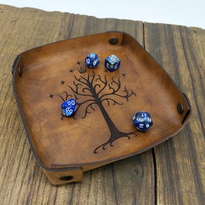 Può includere: Un vassoio per dadi quadrato in pelle marrone con un disegno ad albero e quattro dadi blu. Il vassoio ha un bordo rialzato e bottoni automatici in metallo ad ogni angolo. I dadi sono blu con numeri bianchi.