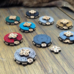 Può includere: Una collezione di segnapunti circolari in legno di vari colori, tra cui rosso, blu, verde, grigio e marrone chiaro. Ogni segnapunti presenta un quadrante rotante centrale con numeri e un pezzo superiore a croce con motivi incisi.