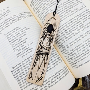 Könnte beinhalten: Holz-Lesezeichen mit einem geschnitzten Design einer Kapuzenfigur. Die Schwarz-Weiß-Illustration zeigt das Gesicht und die Kleidung der Figur im Detail. Das Lesezeichen liegt auf den Seiten eines Buches.