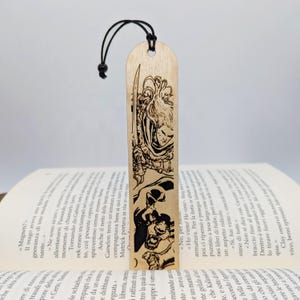 Könnte beinhalten: Holz-Lesezeichen mit einer schwarzen Illustration eines Kriegers und eines Monsters. Das Lesezeichen hat eine schwarze Kordel und befindet sich in einem offenen Buch. Das Lesezeichen ist aus Holz.